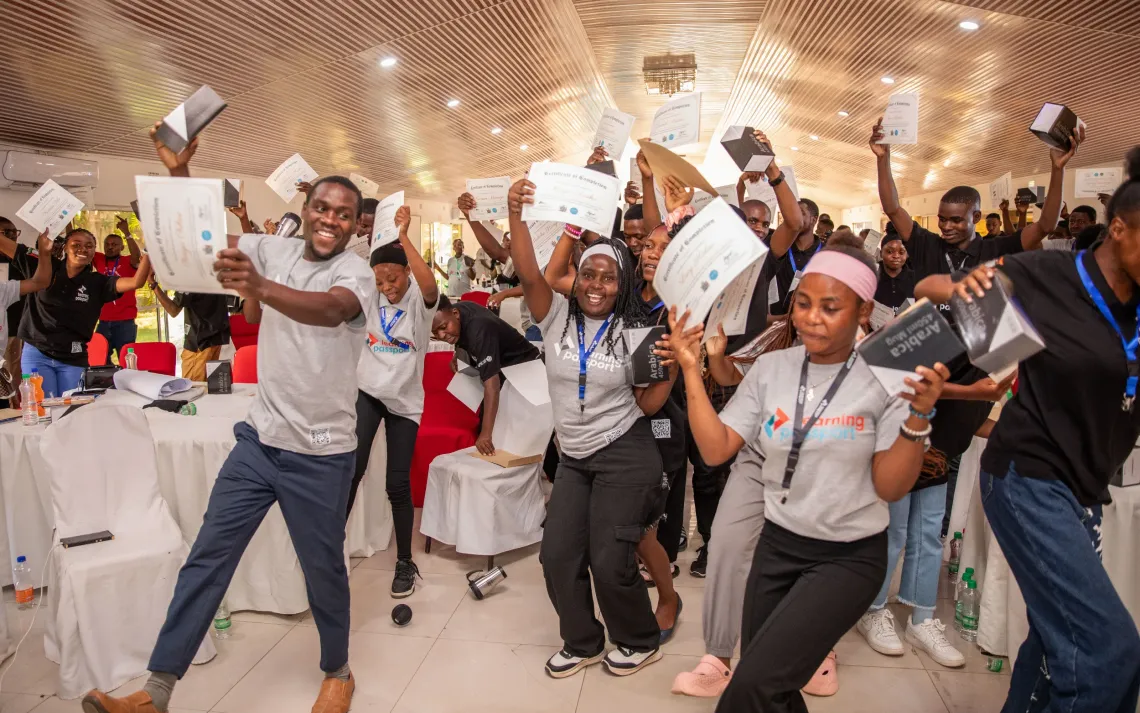 A group of energetic youth participants celebrate at a Learning Passport event, holding up certificates of completion and Airtel-branded tablet boxes. They smile, cheer, and dance together in a brightly lit hall, showing excitement and pride in their achievement.