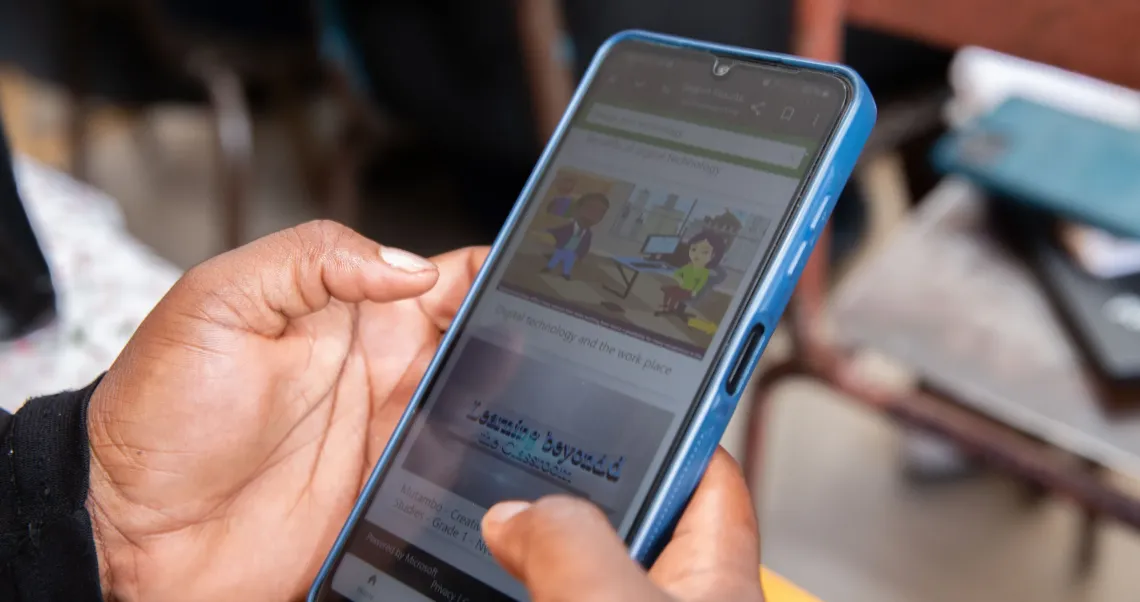A close-up of a student&rsquo;s hand next to a smartphone on a wooden school desk, displaying the Learning Passport app course interface.