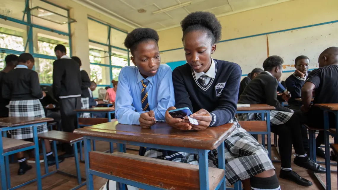 Two students sit at a desk in a classroom, looking at a mobile phone. One student is holding a small white card with login details, likely for accessing the Learning Passport platform. Other students are visible in the background, engaging in similar digital learning activities.
