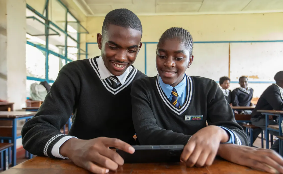 Two smiling secondary school students pose for the camera at a desk with a smartphone in hand, wearing school uniforms&mdash;one with an Academic Prefect badge.
