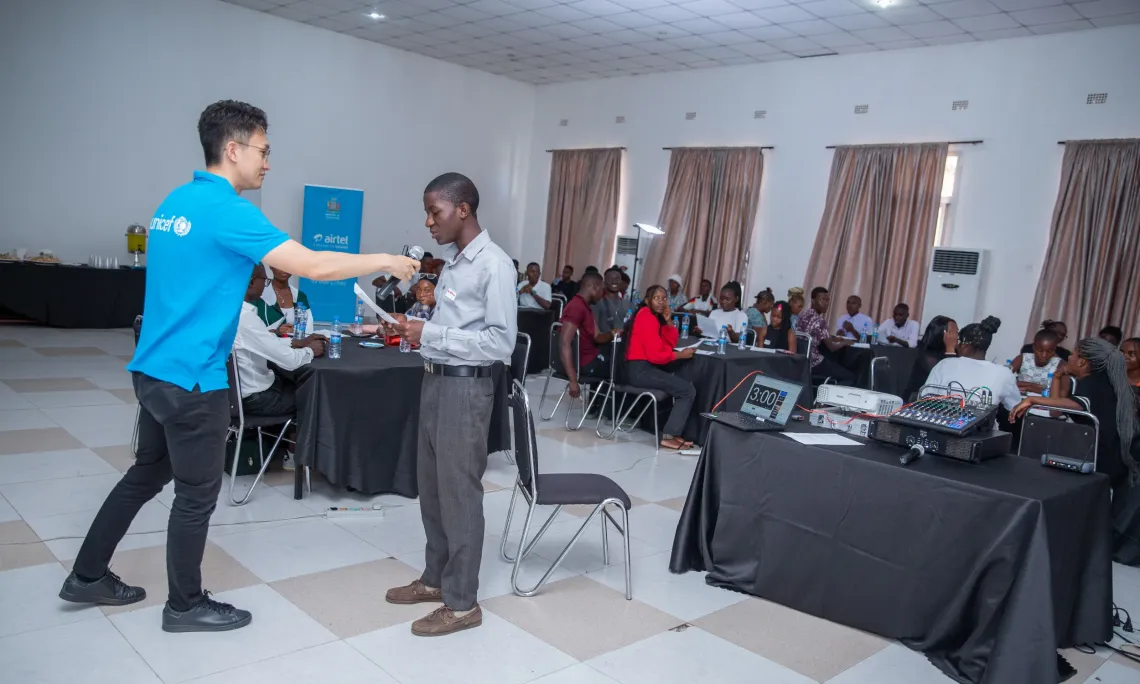 UNICEF Zambia Education Officer hands the microphone to a youth advocate during a group presentation at the final Learning Passport Youth Advocacy Programme meeting. The room is full of engaged participants seated at round tables, with banners in the background showing partners including the Ministry of Education and Airtel.