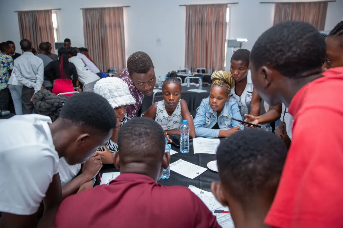 A group of young people gather closely around a table during a workshop activity. They appear engaged and focused, reading from printed materials and discussing something as a team. The setting is a large meeting room with several tables and participants in the background.