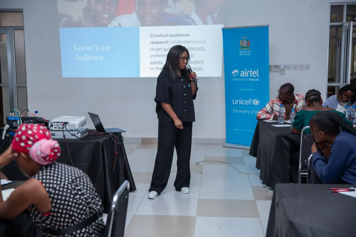 Youth advocates sit attentively around tables during a content creation session, as a UNICEF Zambia team member delivers a presentation. A banner behind her features the logos of the Ministry of Education, Airtel Zambia, and UNICEF.