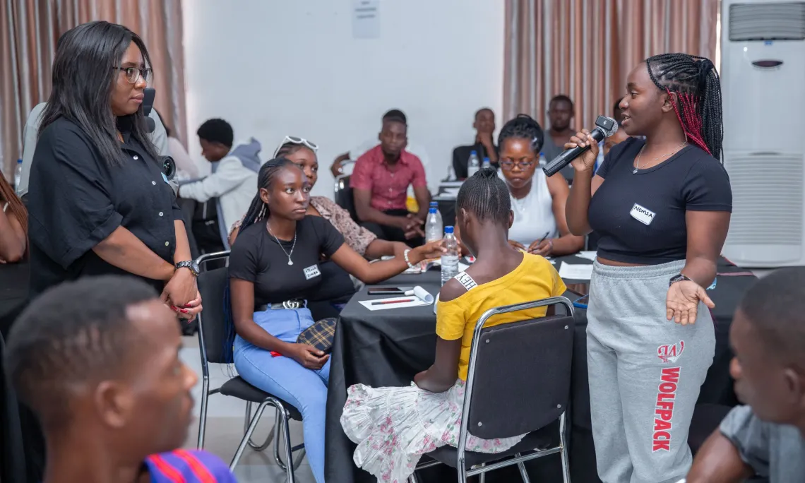 A young woman stands and speaks into a microphone during a group workshop, while a facilitator listens attentively. Other youths sit around tables, watching and participating in the discussion. The setting appears to be an indoor training or dialogue session, with notebooks, water bottles, and name tags visible on the tables.