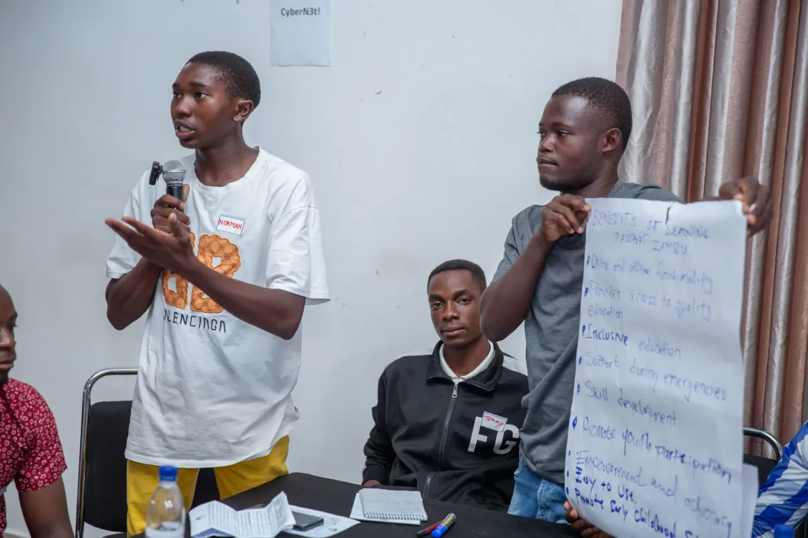 A youth advocate stands and speaks confidently into a microphone while another holds a large handwritten poster outlining the benefits of the Learning Passport Zambia platform, including online access to quality education and youth participation.
