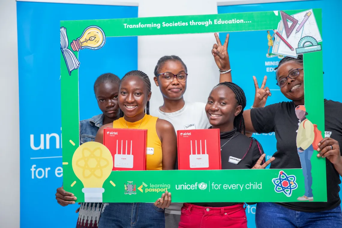 A group of smiling youth advocates pose together holding Airtel-branded routers and framed within a colorful UNICEF-branded photo frame that reads &ldquo;Transforming Societies through Education.&rdquo; The backdrop features Ministry of Education and UNICEF banners.