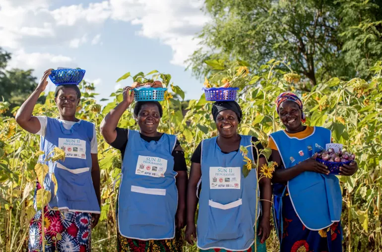 Four smiling women farmers wearing blue SUN II Programme aprons stand in a sunflower field, proudly holding baskets of fresh produce such as sweet potatoes, onions, and greens. Three balance the baskets on their heads while one holds hers in her hands. The scene is bright and sunny, capturing a moment of joy and empowerment.