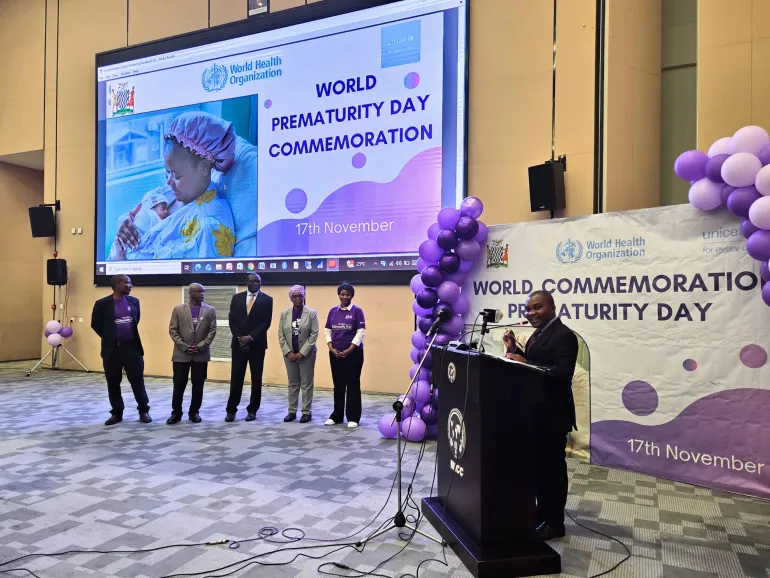 A speaker stands at a podium addressing an audience during a World Prematurity Day commemoration event held on 17th November. Behind him, a large screen displays the event banner featuring the World Health Organization, UNICEF, and Government of Zambia logos, along with an image of a mother holding a premature baby. To the left, six officials stand in a row wearing formal attire and purple event T-shirts. The backdrop and stage decorations include purple and white balloons, symbolizing World Prematurity Day