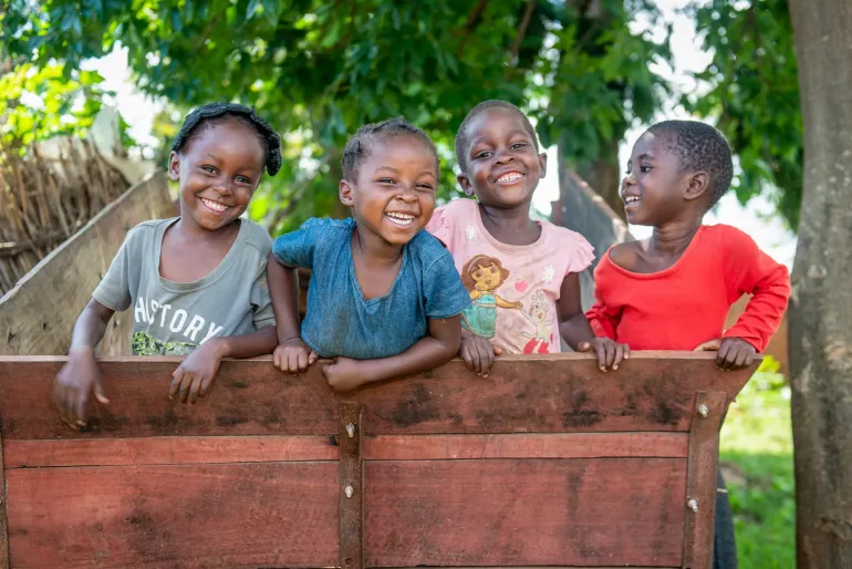 Four young Zambian children smiling and laughing together while leaning over a wooden fence, with green trees in the background. Their joyful expressions capture a sense of childhood happiness and community.